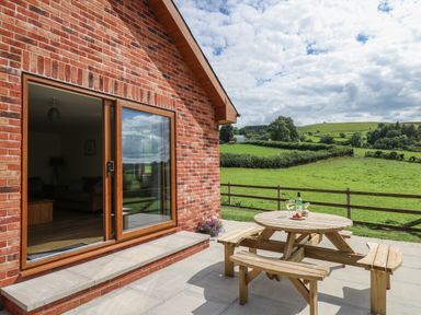 A patio with a wooden picnic table and wine glasses outside a brick house with countryside fields at Awel y Bryn in Rhayader
