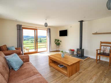 A living room with brown sofas and a wooden coffee table near a wood stove and sliding glass doors at Awel y Bryn in Rhayader