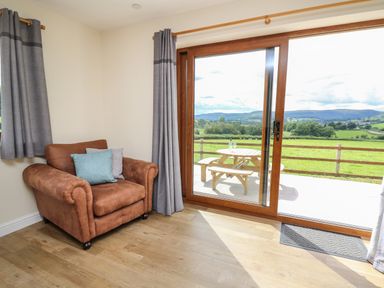 A room with a brown armchair near large glass sliding doors showing an outdoor picnic table and a green landscape at Awel y Bryn in Rhayader