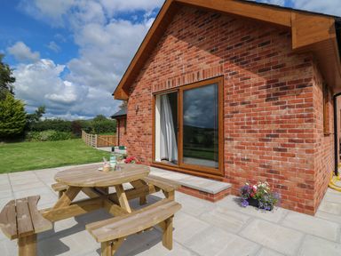 A patio with a wooden picnic table and benches outside a brick house with sliding glass doors at Awel y Bryn in Rhayader