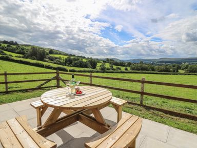 A wooden picnic table on a patio with a bottle of wine two glasses and a plate of tomatoes overlooking green fields and hills at Awel y Bryn in Rhayader