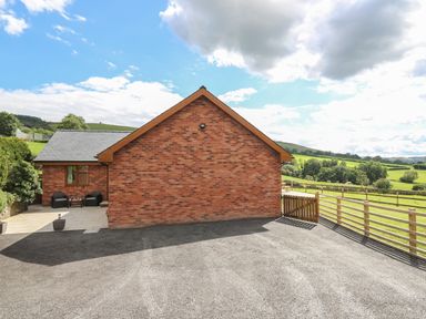 A brick house with a paved patio and outdoor chairs next to a gravel driveway with wooden fencing and fields in the background at Awel y Bryn in Rhayader