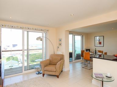 A living area with an armchair and floor lamp next to large sliding glass doors and a dining area with a glass table and four chairs at Sunset View in Castletown Portland
