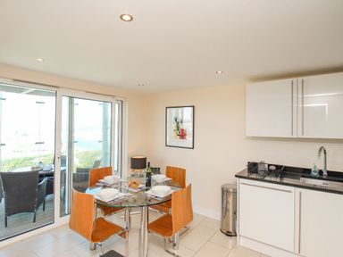 A dining area with a glass table set for four next to a kitchen with white cabinets and a balcony with outdoor seating at Sunset View in Castletown Portland