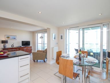 A dining area with a glass table set for four next to sliding glass doors and an open living room with a TV and chair at Sunset View in Castletown Portland