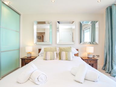 A bedroom with a bed with white linens pillows and rolled towels mirrors above side tables with lamps and blue curtains at Sunset View in Castletown Portland