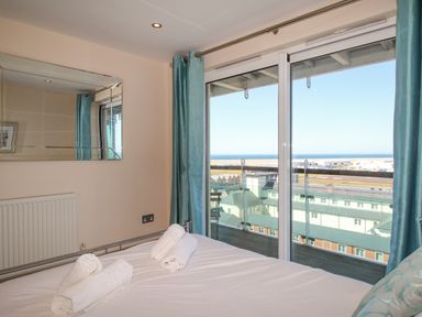 A bedroom with a bed and rolled towels in front of a window with blue curtains overlooking buildings and the sea at Sunset View in Castletown Portland