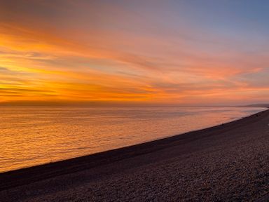A sunset over a calm sea with a pebble beach and a few people along the shore at Sunset View in Castletown Portland