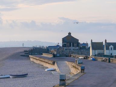 A pebbled beach with small boats near a concrete promenade and houses in the background at Sunset View in Castletown Portland