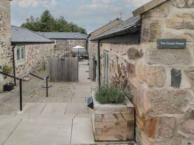 An outdoor stone walkway between buildings with a wooden fence and patio furniture at The Coach House in Bamburgh