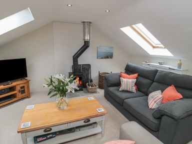 A living room with a gray sofa and wooden coffee table with flowers by a wood burning stove at The Coach House in Bamburgh