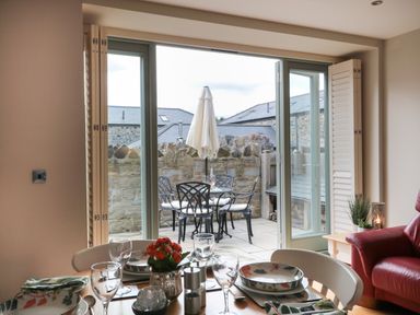 A dining table set with plates glasses and a flower pot next to a red armchair overlooking a patio with an outdoor table chairs and umbrella at The Coach House in Bamburgh