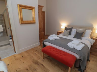 A bedroom with a bed, bedside tables with lamps, a red bench, folded towels, a wooden floor, and a gold framed mirror on the wall at The Coach House in Bamburgh