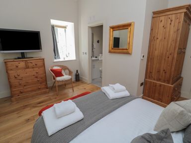 A bedroom with a bed with folded towels a wooden dresser a wicker chair with a cushion a wall mirror and wooden wardrobe at The Coach House in Bamburgh