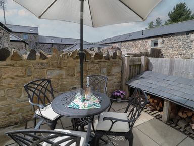 An outdoor patio with a round metal table and four chairs under a large umbrella near a stone wall and wood storage at The Coach House in Bamburgh