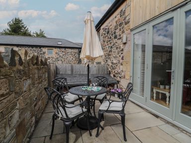 A stone walled patio with a round metal table and four cushioned chairs and a closed umbrella at The Coach House in Bamburgh