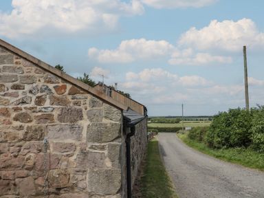 A stone building next to a gravel road with fields and bushes in the background at The Coach House in Bamburgh