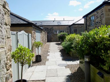A stone pathway between stone buildings with potted plants and bushes at The Coach House in Bamburgh
