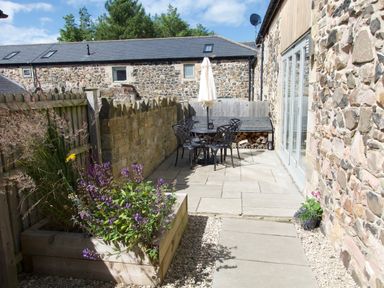 An outdoor patio area with a table and chairs and a closed umbrella next to a stone building at The Coach House in Bamburgh