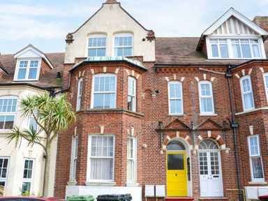 A row of brick townhouses with white framed windows and doors with one door painted yellow with a tree and bins in front at Flat 3 in Cromer