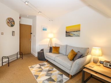 A living room with a grey sofa with cushions a wooden side table with a lamp and radio a geometric rug wall art and a wooden door at Flat 3 in Cromer