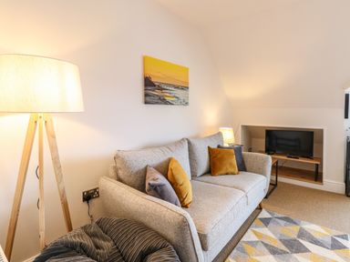 A living room with a grey sofa and cushions a floor lamp a small table with a lamp a television on a low wooden shelf and a geometric patterned rug at Flat 3 in Cromer