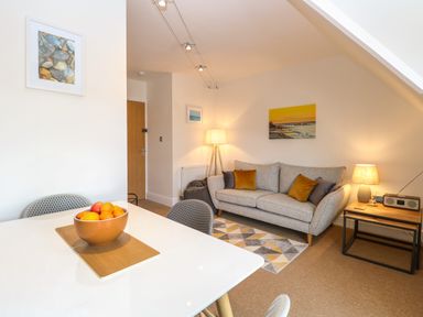 A living room with a gray sofa with pillows a wooden side table with a lamp and radio a bowl of fruit on a white table at Flat 3 in Cromer