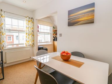 A dining area with a white table and four chairs a bowl of fruit on the table curtains on windows and a beach painting on the wall at Flat 3 in Cromer