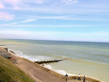 A coastal pathway with two people fishing by a sea wall and wooden groynes in the water at Flat 3 in Cromer