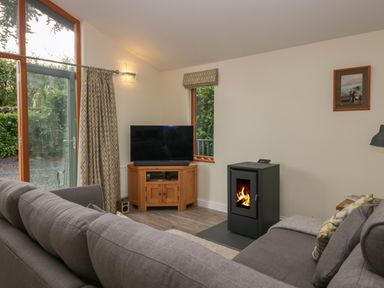 A living room with a gray sofa a wood burning stove a wooden TV stand with a television and windows showing greenery at Bay Tree Lodge in Cartmel
