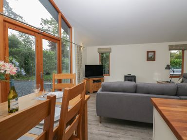 A dining table with chairs and wine in a living room with a gray sofa television and windows at Bay Tree Lodge in Cartmel