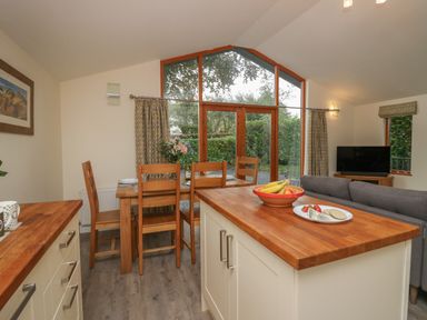 A kitchen and dining area with a wooden dining table and chairs next to large windows and a kitchen island with fruit at Bay Tree Lodge in Cartmel