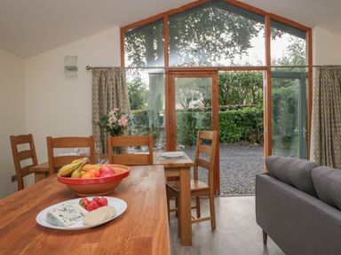 A dining area with wooden tables and chairs a bowl of fruit and a plate with cheese strawberries and crackers with glass doors leading outside at Bay Tree Lodge in Cartmel