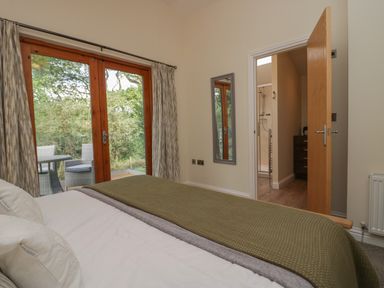 A bedroom with a bed facing glass doors leading to a balcony with outdoor furniture and an open door to a bathroom at Bay Tree Lodge in Cartmel