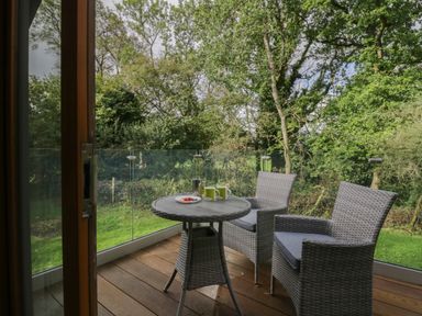 A balcony with a round table and two wicker chairs overlooking trees and grass at Bay Tree Lodge in Cartmel