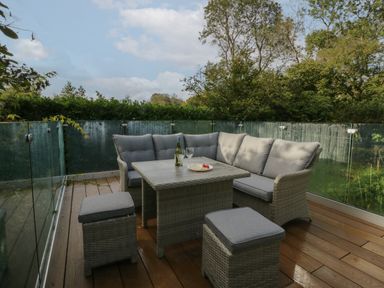 An outdoor patio with grey cushioned seating a table with a bottle and glasses surrounded by glass railing at Bay Tree Lodge in Cartmel