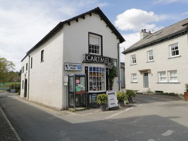 A street corner with a white building labeled Cartmel village shop and a phone booth near a road sign at Bay Tree Lodge in Cartmel