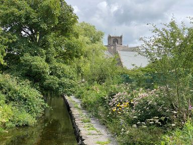 A narrow river beside a gravel path with bushes flowers and trees with a stone building with a clock tower in the background at Bay Tree Lodge in Cartmel
