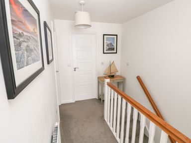 A hallway with white walls and carpeted floor featuring framed pictures on the wall a small table with a model sailboat and a door at the end near stairs with a wooden handrail at Ty Glas in Ogmore-by-Sea near St Brides Major
