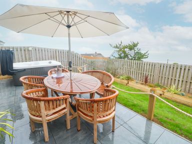 An outdoor patio with a round wooden table and chairs under a large white umbrella near a hot tub at Ty Glas in Ogmore-by-Sea near St Brides Major