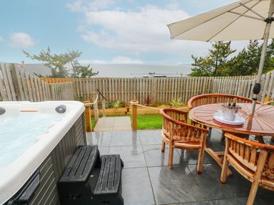 An outdoor patio with a hot tub on the left and a wooden round table with four chairs and an umbrella on the right at Ty Glas in Ogmore-by-Sea near St Brides Major