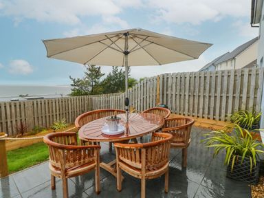 An outdoor patio with a wooden table and six chairs under a large beige umbrella at Ty Glas in Ogmore-by-Sea near St Brides Major