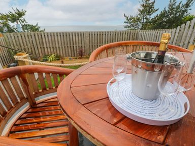 A wooden outdoor table with three champagne glasses and a bucket with a bottle overlooking a fenced garden and sea view at Ty Glas in Ogmore-by-Sea near St Brides Major