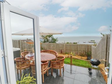 A patio with a round wooden table and chairs a barbecue and umbrella overlooking a fenced garden and sea at Ty Glas in Ogmore-by-Sea near St Brides Major