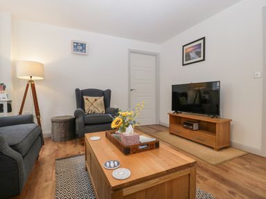A living room with a wooden coffee table with a tray of flowers a grey armchair a floor lamp a television on a wooden stand a framed picture on the wall at Ty Glas in Ogmore-by-Sea near St Brides Major
