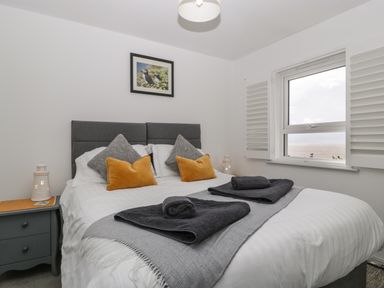 A bedroom with a double bed, gray and orange pillows, gray towels on the bed, a side table with a decorative lighthouse, and a window with white shutters at Ty Glas in Ogmore-by-Sea near St Brides Major