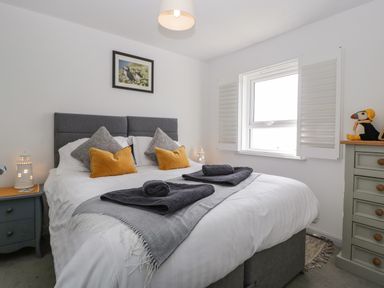 A bedroom with a double bed with gray headboard and pillows and yellow cushions two rolled towels on a gray runner bedside cabinets with lighthouse lamps and a chest of drawers with a puffin stuffed toy near a window with white shutters at Ty Glas in Ogmore-by-Sea near St Brides Major