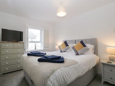 A bedroom with a bed, bedside table with lamp, chest of drawers with TV, and a window with shutters at Ty Glas in Ogmore-by-Sea near St Brides Major