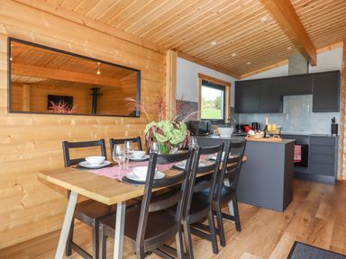 A dining area with a wooden table and black chairs next to a kitchen with dark cabinets and wooden ceiling at Foxglove Lodge in Hittisleigh