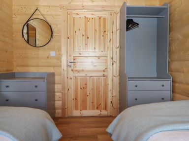 A bedroom with wooden walls a wooden door two gray dressers a round wall mirror and two beds with light blankets at Foxglove Lodge in Hittisleigh